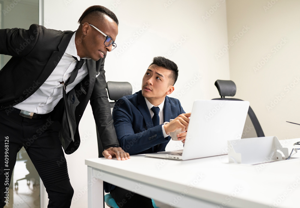 Serious young Asian male boss in formal suit with cup of coffee pointing at laptop screen and asking African American manager to explain problem solution during meeting in office Serious young Asian male boss in formal suit with cup of coffee pointing at laptop screen and asking African American manager to explain problem solution during meeting in office