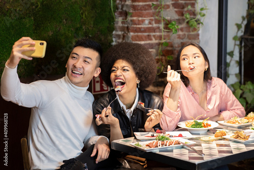 Group of positive young multiracial friends having fun and taking selfie on smartphone while enjoying delicious meal together in cozy cafe