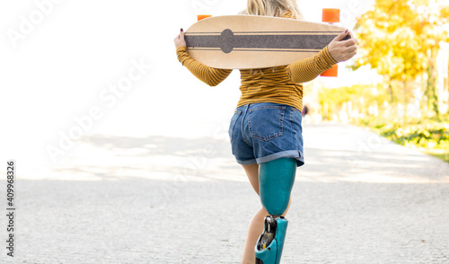 Back view of crop anonymous female skater with bionic prosthesis standing in city with longboard and enjoying summer