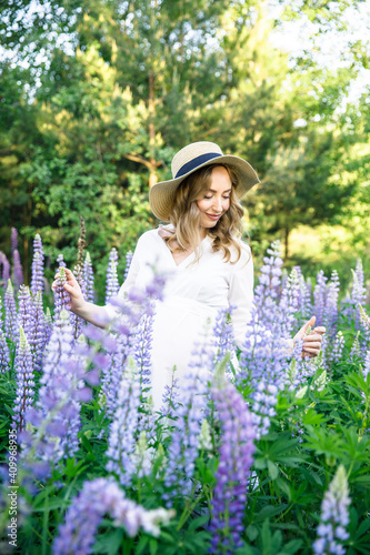 young beautiful pregnant woman in a white dress and hat walks across the field with lupins