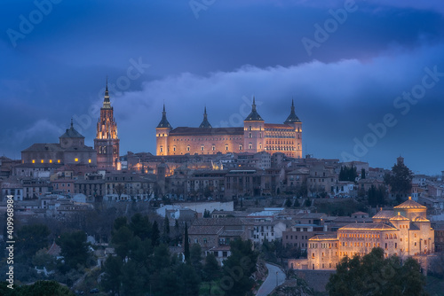 Aerial view of old city with historic buildings and stone castle on hill against cloudy sky during sunset in Toledo, Spain