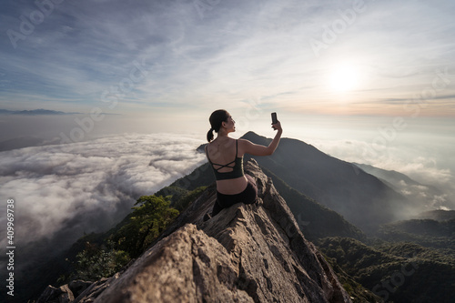 Back view of female ethnic hiker sitting on top of Yuanzui Mountain and taking self portrait on smartphone photo