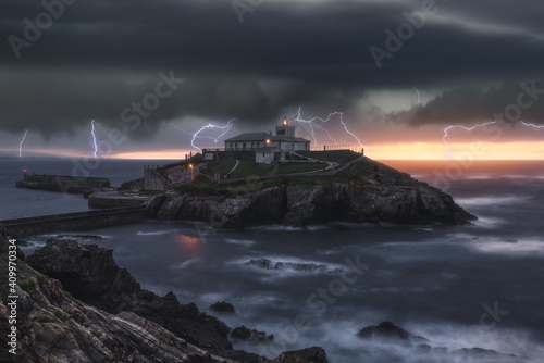 Picturesque view of thunderstorm with flashes of lightnings above lonely house on island in stormy sea at sunset photo