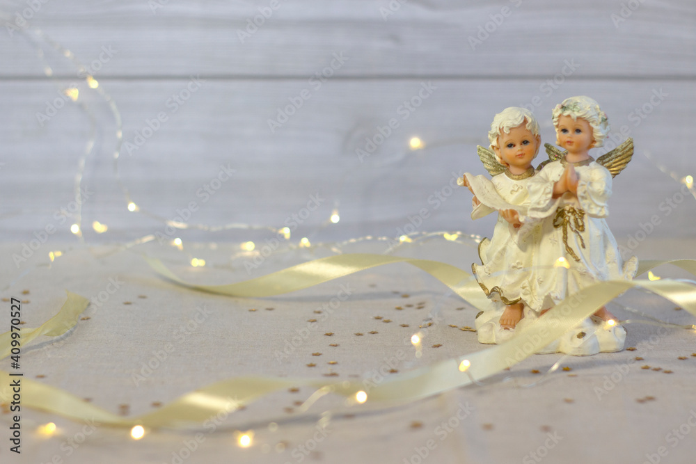 Little ceramic angels pray and sing songs, on a blurred background, satin ribbon and lights