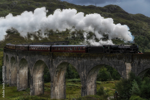 Old fashioned steam train riding along Glenfinnan Viaduct located in mountains on cloudy day in Scotland
