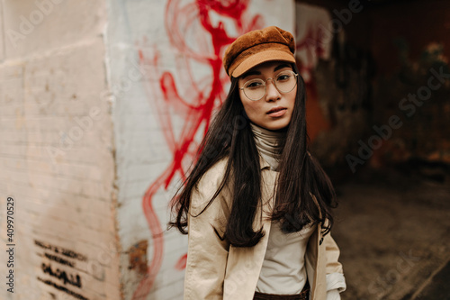 Brunette woman in glasses, cap and trench coat looks into camera amid abandoned room