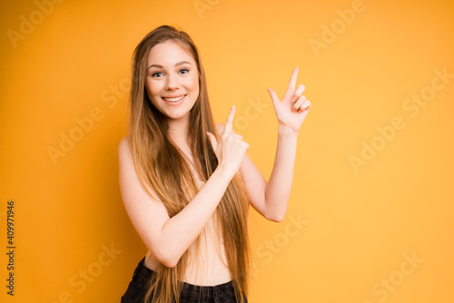 A cute caucasian girl with long hair in a beige top and black jeans smiles against an orange background and points her fingers to the right. space for text