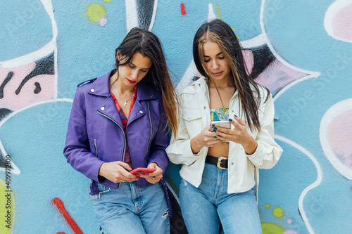 Anti-social trendy millennial women in stylish wear standing near colorful painted wall browsing on smartphone