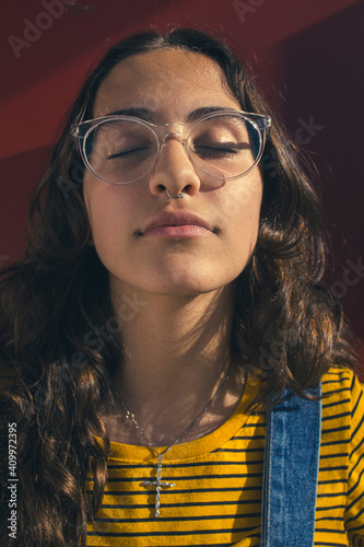 Close up portrait of a thoughtful and introspective teenager girl wearing clear transparent glasses and colorful clothes against red wall with eyes shut.