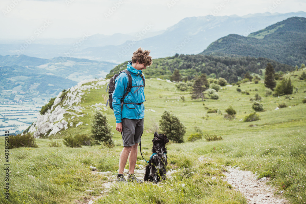 A young male standing with his dog in the beautiful mountain landscape and looking each other. Recreation and healthy lifestyle outdoors, summer in mountains.