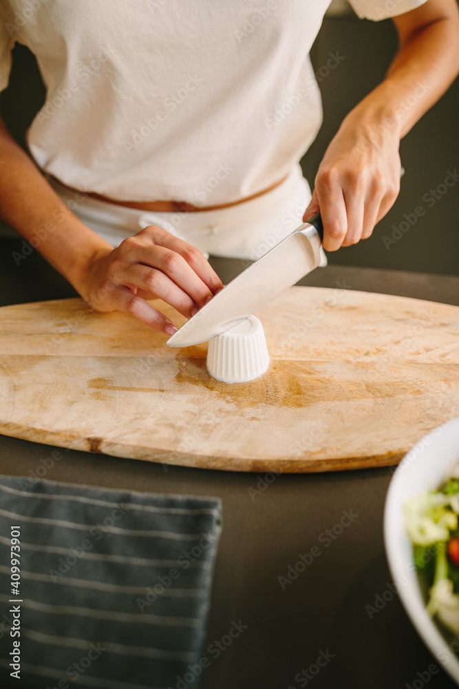 Unrecognizable delighted female cutting Feta cheese on chopping board while preparing ingredients for delicious homemade vegetable salad Unrecognizable delighted female cutting Feta cheese on chopping board while preparing ingredients for delicious homemade vegetable salad