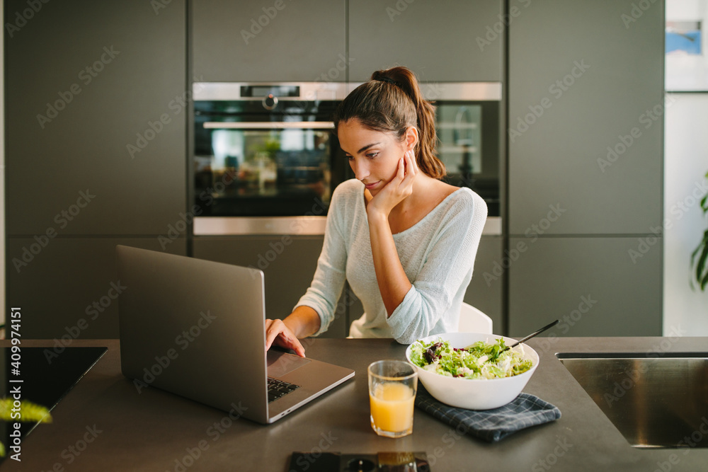 Female freelancer using laptop and working on project online while sitting at table with glass of juice and bowl of salad in kitchen Female freelancer using laptop and working on project online while sitting at table with glass of juice and bowl of salad in kitchen