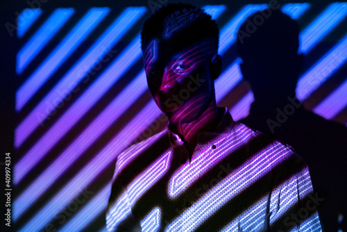 Unemotional African American male standing in neon light in shape of stripes in dark studio and looking at camera