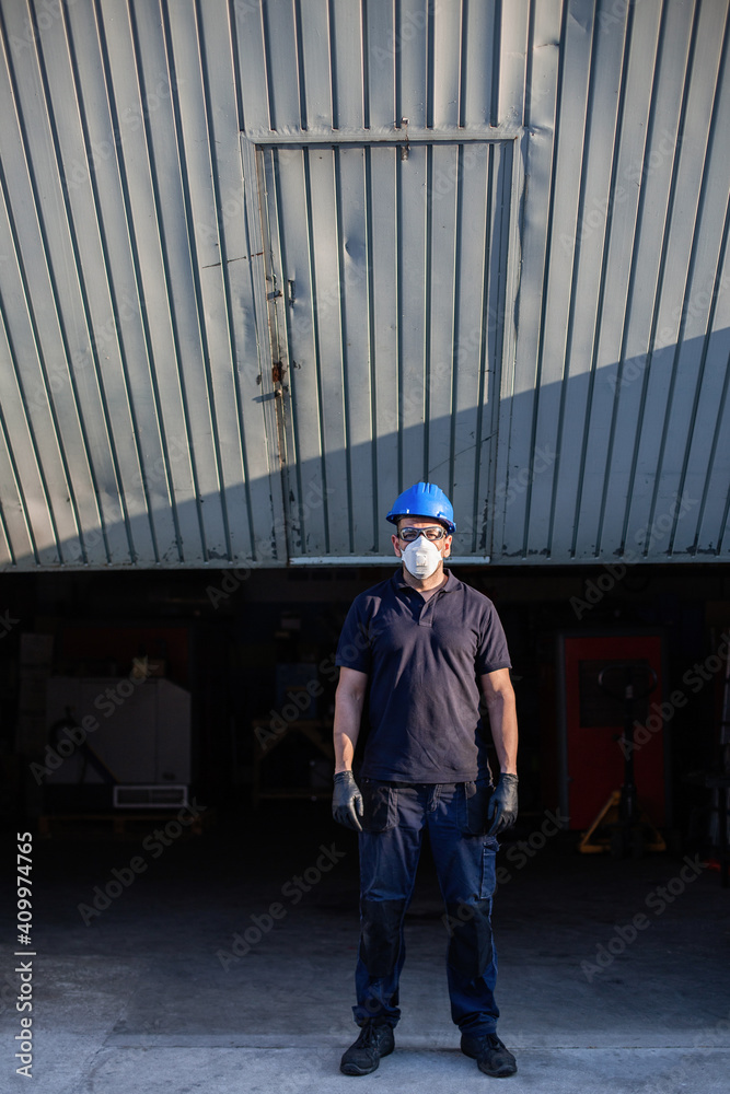 Adult male mechanic in protective glasses and respirator with blue hardhat looking at camera while standing near workshop building on street Adult male mechanic in protective glasses and respirator with blue hardhat looking at camera while standing near workshop building on street
