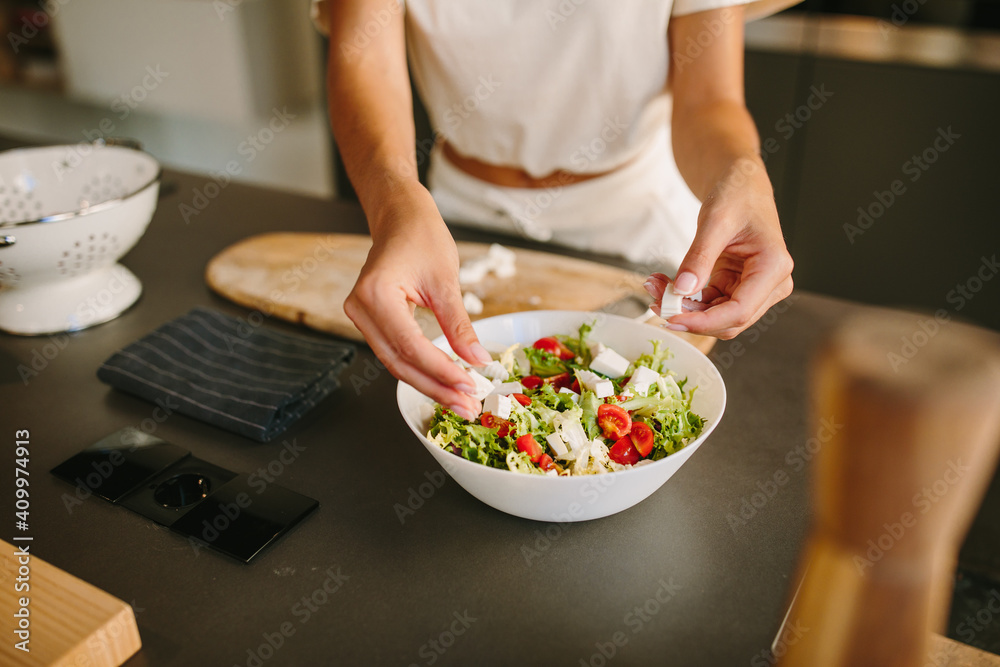 Unrecognizable female adding Feta cheese in fresh vegetable salad while preparing healthy nutritious lunch in kitchen Unrecognizable female adding Feta cheese in fresh vegetable salad while preparing healthy nutritious lunch in kitchen