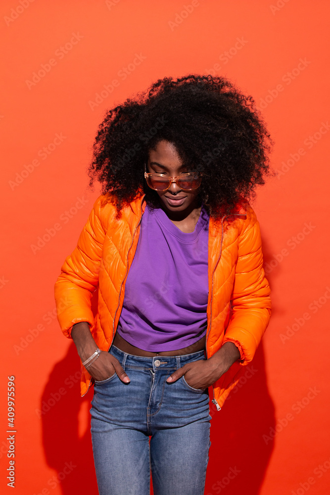 Young black female in purple shirt with denim and orange jacket standing with hands in pockets on red background looking down Young black female in purple shirt with denim and orange jacket standing with hands in pockets on red background looking down