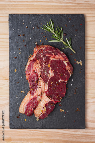 Top view of fresh uncooked rib eye beef steak with rosemary sprig and dried spices  on black slate board