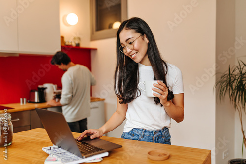 Brunette woman in t-shirt and jeans is looking into laptop and holding white cup © Look!