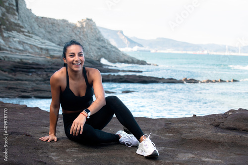 Delighted fit female in activewear sitting on beach on background of amazing seascape and looking at camera after workout