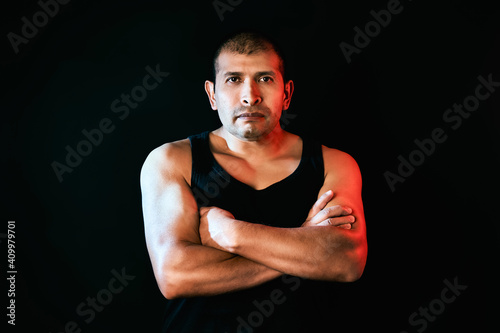 Fighter and strong Hispanic man wears a t-shirt that shows his muscular crossed arms and looks at the camera seriously. Isolated on black background