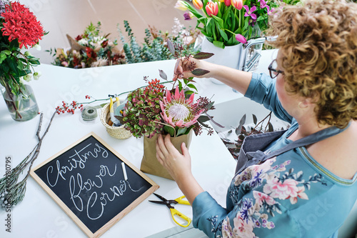 From above of female florist in apron standing in shop and creating bouquet from Hydrangea and King protea flowers photo