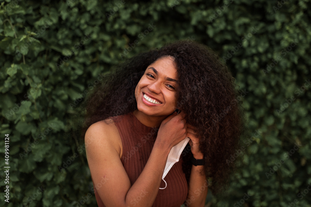 Young black female in protective mask and with curly hair standing in urban park and looking at camera during coronavirus epidemic Young black female in protective mask and with curly hair standing in urban park and looking at camera during coronavirus epidemic