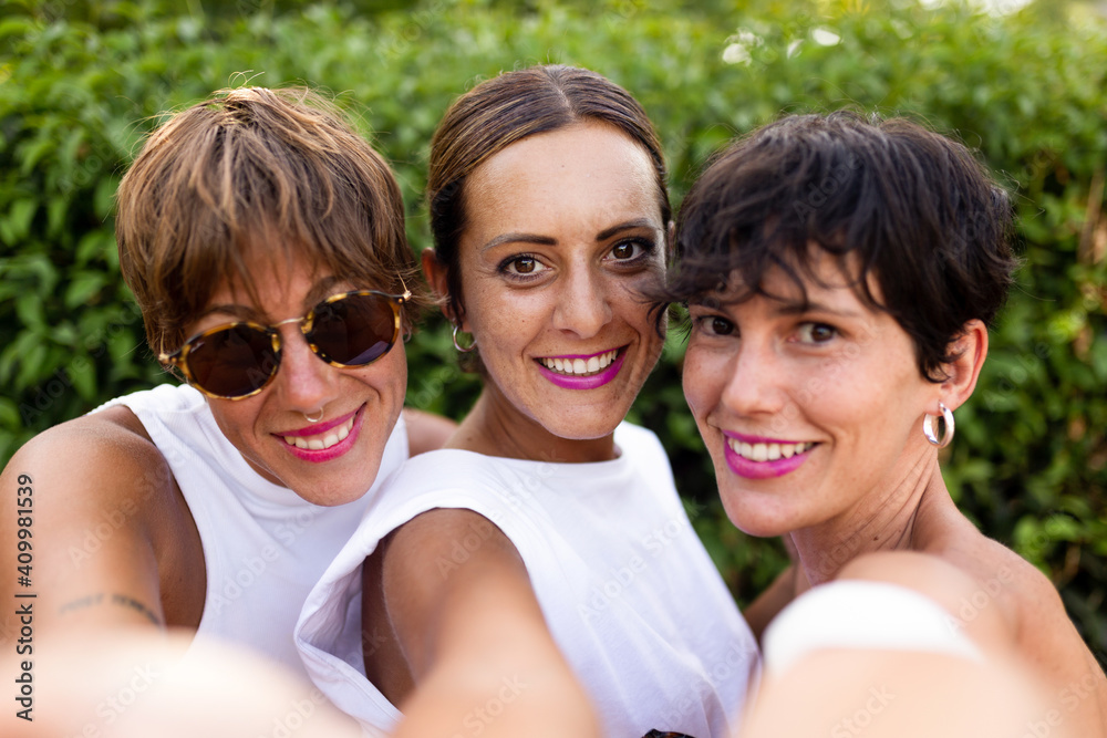 Company of adult female friends standing in summer park and taking self portrait while charmingly smiling and looking at camera Company of adult female friends standing in summer park and taking self portrait while charmingly smiling and looking at camera