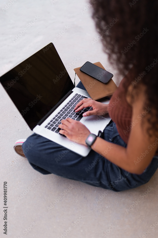 African American self employed woman sitting on street with laptop and browsing smartphone African American self employed woman sitting on street with laptop and browsing smartphone