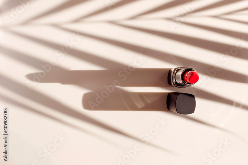 Red lipstick sun shadows from palm leaf on beige background flat lay top view copy space. Beauty and cosmetics background. Decorative cosmetics, makeup, women's lipstick, beauty brand product design