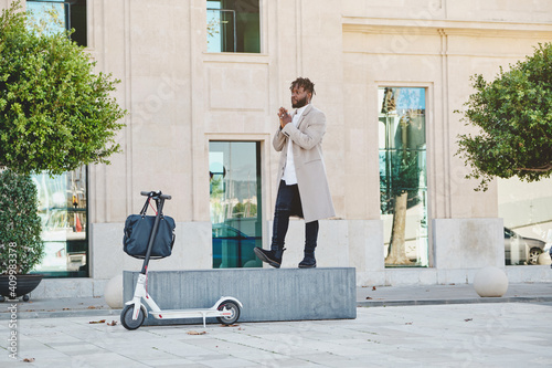 Side view of african American guy in casual outfit standing on the street with electric scooter in city