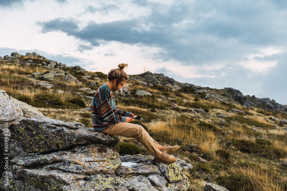 Side view of female tourist sitting on rough rock and using smartphone while relaxing during trekking in mountains Side view of female tourist sitting on rough rock and using smartphone while relaxing during trekking in mountains