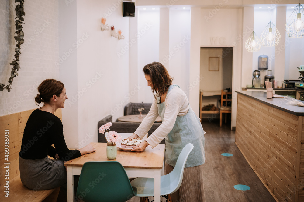 Smiling female small business owner serving delicious cupcake to customer sitting at table in cozy confectionery cafe Smiling female small business owner serving delicious cupcake to customer sitting at table in cozy confectionery cafe