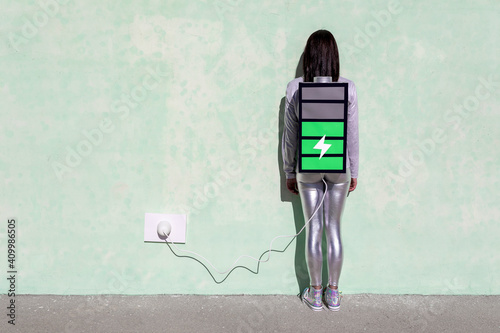 Back view of unrecognizable female with long dark hair in stylish outfit standing on street near wall with battery shaped backpack connected to charger
