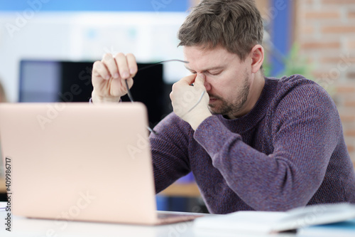 Фотография Tired man siting in front of laptop screen and holding glasses in hands