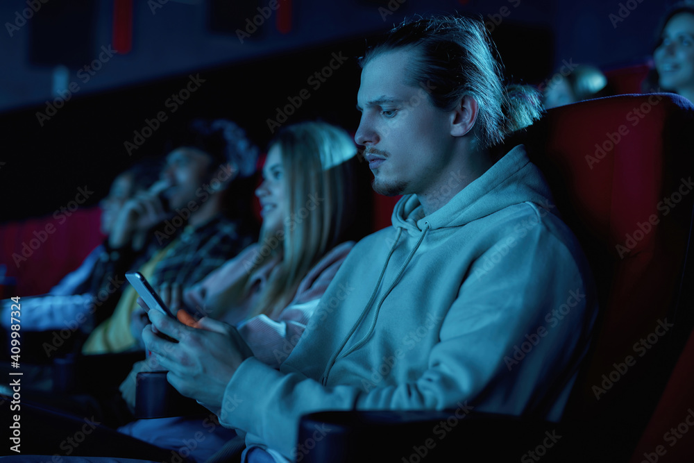 Focused young guy using his smartphone while missing boring movie at the cinema