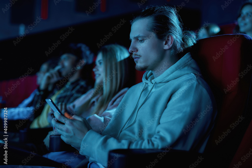 Fototapeta premium Focused young guy using his smartphone while missing boring movie at the cinema