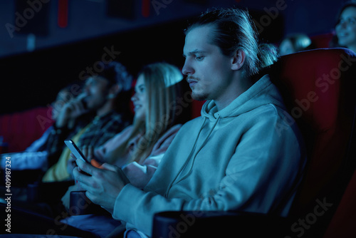 Focused young guy using his smartphone while missing boring movie at the cinema