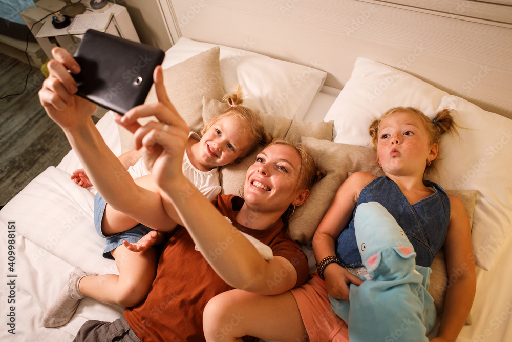 Fototapeta premium Mom and her two daughters are taking selfie on the smartphone relaxing on bed.