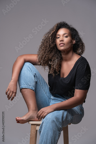 Plus size barefoot young African American female with curly hair dressed in jeans and black shirt sitting on chair and looking at camera against gray background