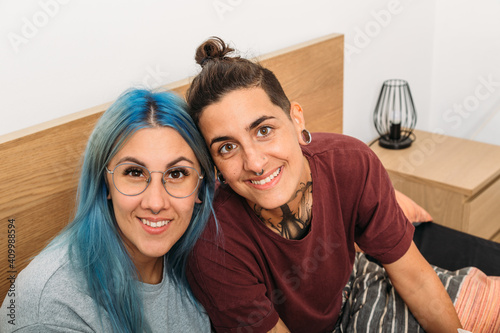 From above tattooed androgynous woman sitting with happy girlfriend with dyed hair resting on bed at home together looking at camera