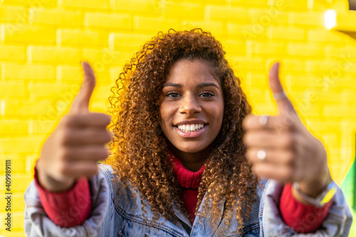 Optimistic young black female millennial with curly hair in stylish clothes showing thumbs up gesture and smiling while looking at camera against yellow wall photo