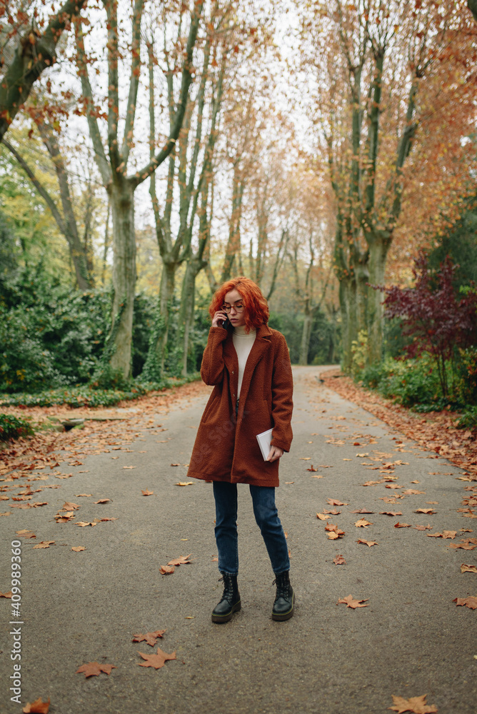 Optimistic female with red hair and in autumn coat standing on alley in park and enjoying smartphone conversation with friend Optimistic female with red hair and in autumn coat standing on alley in park and enjoying smartphone conversation with friend