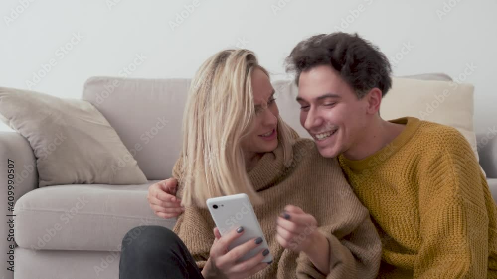 Young happy couple having fun using smartphone at home. Girl and guy talking, laughing, looking at mobile phone buying retail products shopping online, ordering delivery, checking social media apps.