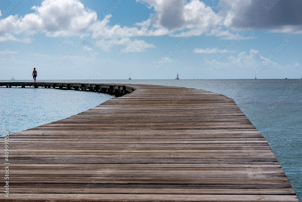 an isolated man walking on a curved wooden pier under a blue cloudy sky. some sailing boats on the calm sea