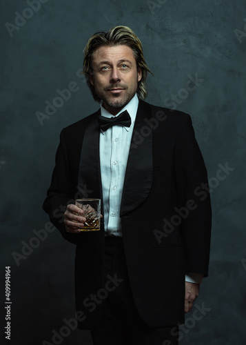 Happy blonde man with stubble beard in tuxedo holding glass of whisky.