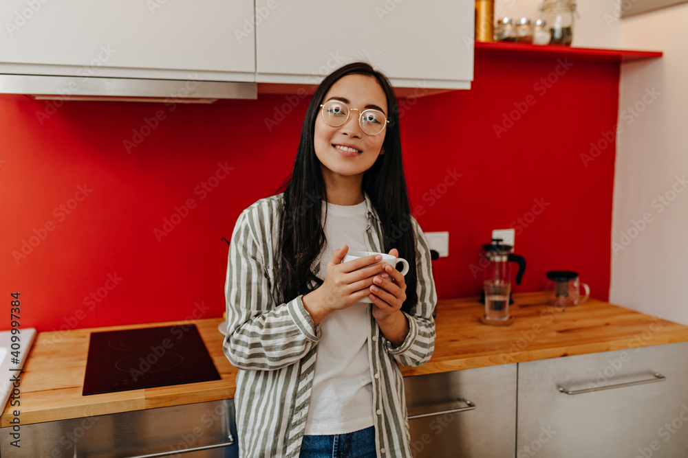 Beautiful Asian girl in striped shirt and glasses looks into camera and holds cup Beautiful Asian girl in striped shirt and glasses looks into camera and holds cup