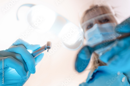 Zahnarzt / Zahnärztin mit Bohrer und OP Leuchte aus Sicht des Patienten dicht über dem Mund mit Corona Maske