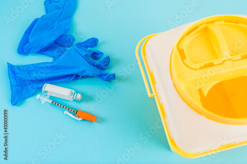 Yellow medical waste container next to blue syringe, vaccine and latex gloves on a blue background
