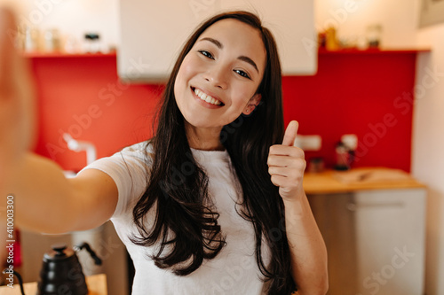 Long-haired woman dressed in white T-shirt makes selfie in kitchen and shows thumb up