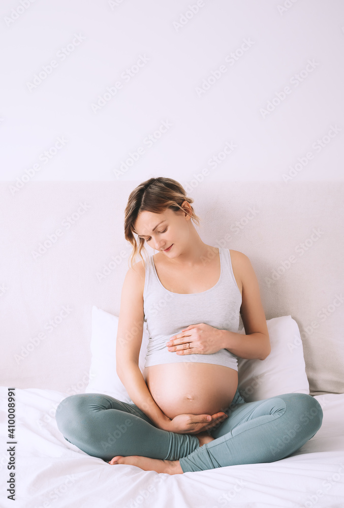 Beautiful relaxed pregnant woman holds hands on her belly in bedroom at home. Concept of wellbeing during pregnancy, maternity, healthcare, gynecology. Beautiful relaxed pregnant woman holds hands on her belly in bedroom at home. Concept of wellbeing during pregnancy, maternity, healthcare, gynecology.